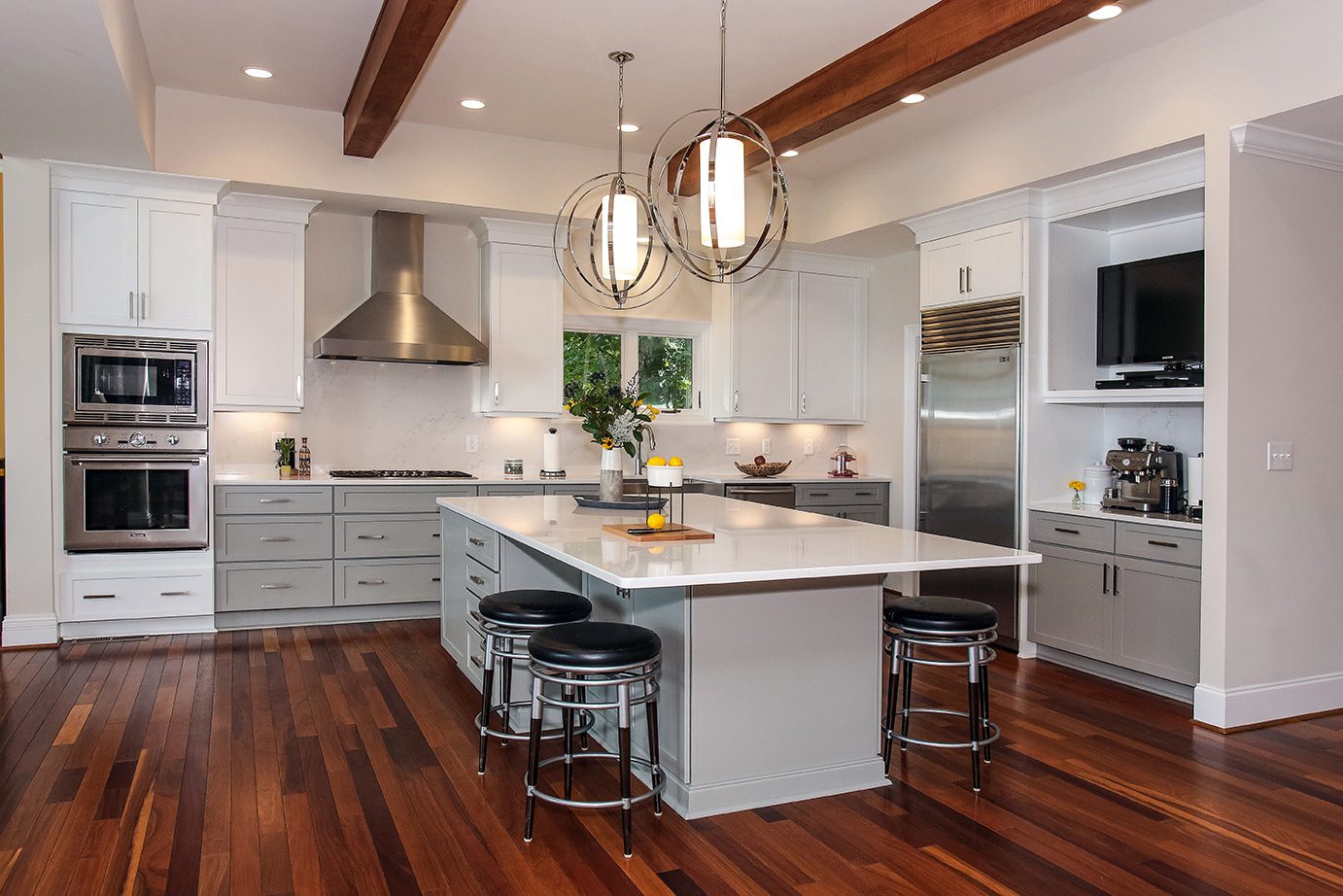 Modern kitchen with white shake style cabinets and island with white granite tops. Brown hard wood floor and exposed beam ceiling. The kitchen has stainless steel appliances and ornate light fixtures over the island and black and silver stools around it